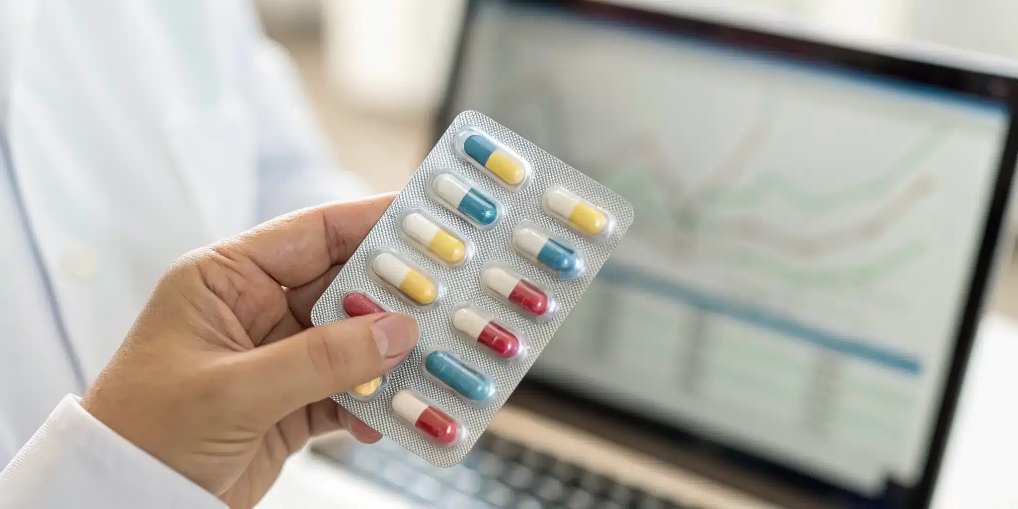 Hand holding pharmaceutical capsules over a laptop with data charts for managing chargebacks.