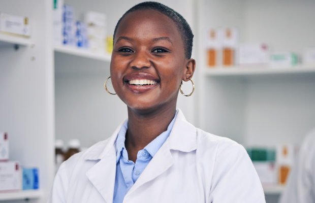 Smiling Black woman in a white lab coat in a pharmacy, looking at the camera with shelves of medicines in the background