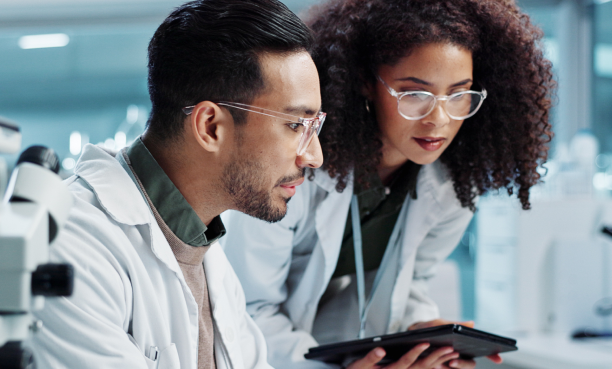 Two scientists in lab coats and safety glasses examine data on a tablet together in a bright lab environment.