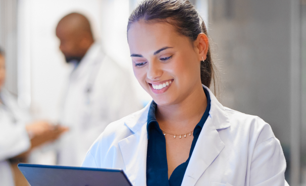 Female scientist in a white lab coat smiles while looking at a tablet in a clinical lab, with colleagues blurred in the background.