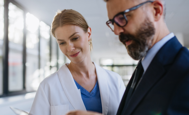 Two healthcare professionals review a document together in a bright clinic hallway; a female clinician in a white coat and blue scrubs smiles slightly as she looks at the paper with a male colleague in a suit and glasses beside her.