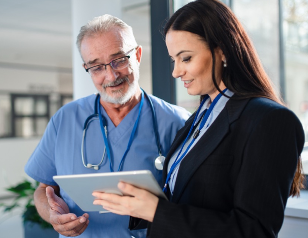 Two healthcare professionals review a tablet together: an older man in blue scrubs with a stethoscope and a woman in a blazer standing by a window in a hospital setting.
